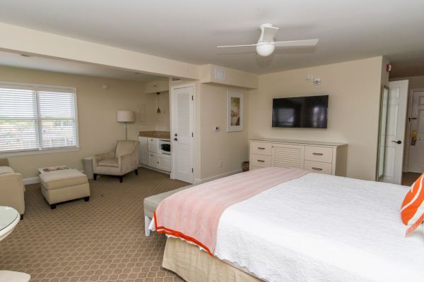 A well-lit hotel room with a bed, chairs, a TV, and some kitchen amenities.